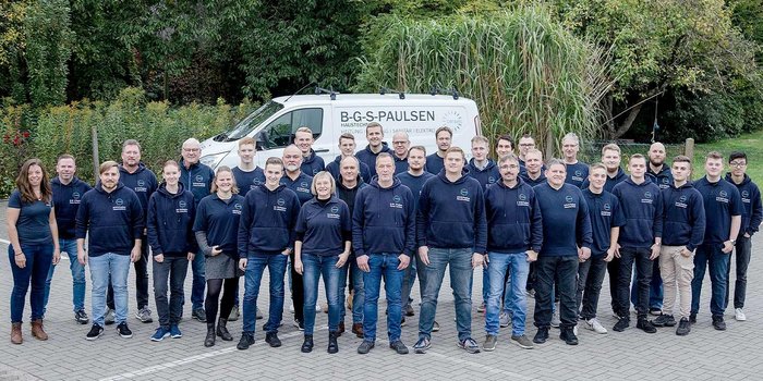 Mitarbeiter eines Unternehmens posieren gemeinsam vor einem weißen Lieferwagen, umgeben von Bäumen und Sträuchern.