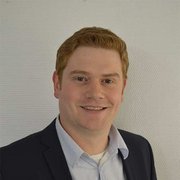 Ein Mann mit roten Haaren lächelt freundlich in einem Anzug vor einer hellen, strukturierten Wand.