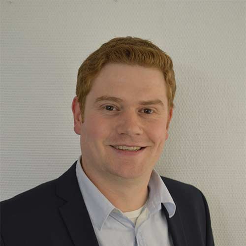 Ein Mann mit roten Haaren lächelt freundlich in einem Anzug vor einer hellen, strukturierten Wand.