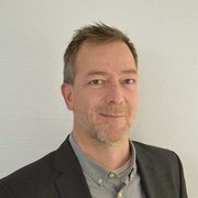 Ein Mann mit kurzen, grauen Haaren und einem leichten Bart lächelt freundlich in die Kamera, gekleidet in einem Anzug.