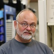 Ein älterer Mann mit Brille und grauem Bart schaut freundlich in die Kamera, umgeben von einem geschäftigen Hintergrund.