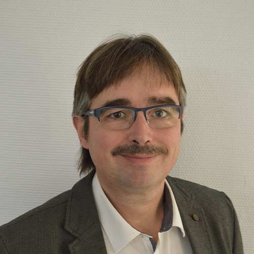 Der Mann hat einen gepflegten Look mit einem Schnurrbart und trägt eine weiße Bluse unter einem grauen Anzug.