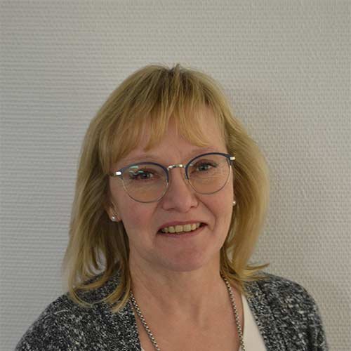 Eine lächelnde Frau mit blonden Haaren und Brille steht vor einer strukturierten weißen Wand.